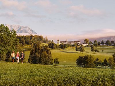 Three hikers walk across a lush green field with distant mountains and a clear blue sky. Trees and a building are visible in the picturesque landscape.