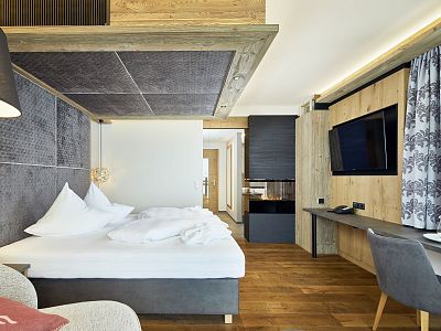 A spacious hotel room with a large bed, white linens, a wall-mounted TV, desk, and wooden decor, featuring a cozy and modern design.