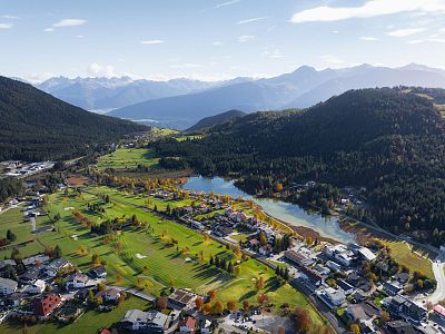 Aerial view of a mountain valley with a lake, village, and green fields surrounded by mountains under a blue sky.