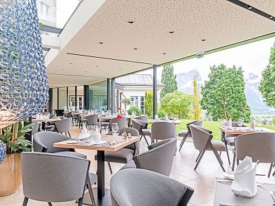 Elegant restaurant with tables and chairs, large windows showcasing greenery and mountain scenery.