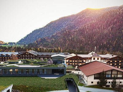 Alpine resort in a mountain valley with traditional architecture and green rooftops, surrounded by forested hills and a clear sky.