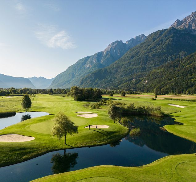 A lush golf course surrounded by mountains, with green fairways, water hazards, and trees under a clear blue sky.