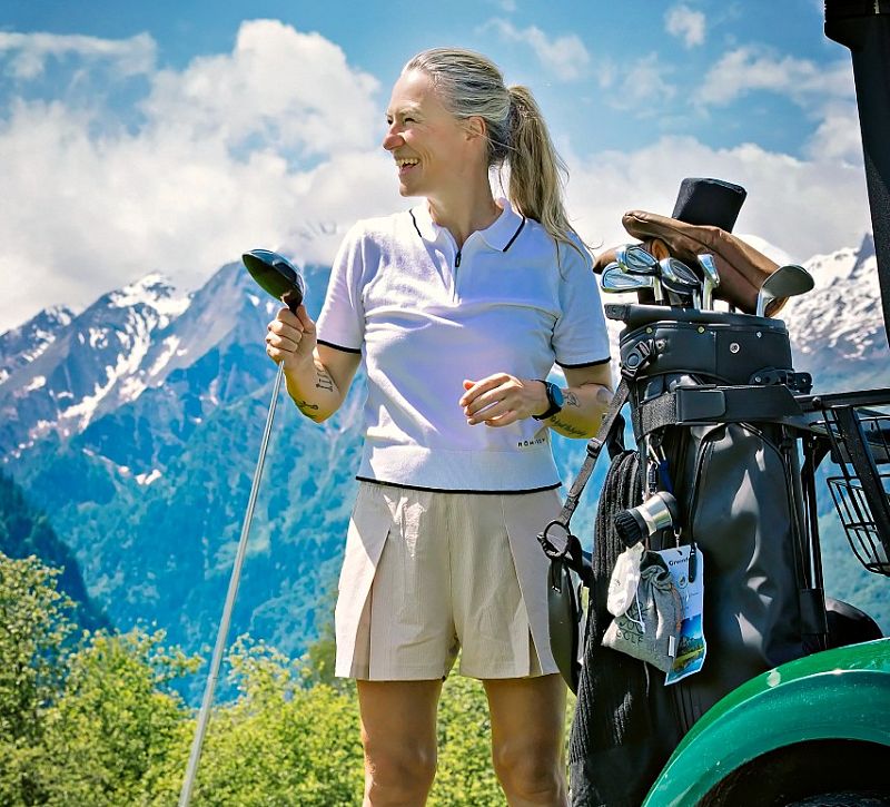 Person golfing with mountains in background, wearing white polo and beige shorts, standing by golf cart with clubs, under a blue sky and snow-covered peaks.