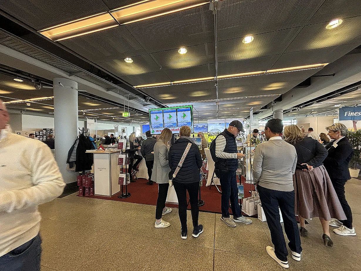 Crowded airport terminal with people standing, monitors displaying information, and various booths and counters visible. A lively travel environment.