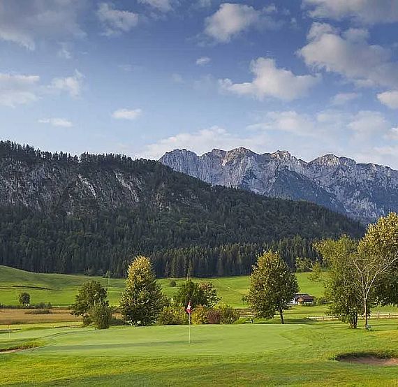 Golf course with lush green grass, trees, and a backdrop of rolling hills and mountains under a partly cloudy blue sky.
