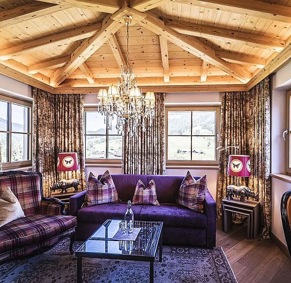 Gemütliches Wohnzimmer mit Holzbalkendecke, lila Sofa und karierten Kissen. Ein Kristalllüster hängt von der Decke, und große Fenster bieten Bergblick.