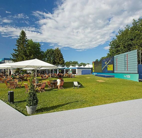 Outdoor space with canopy, tables, chairs on grass, stage on right, trees in background, clear sky.