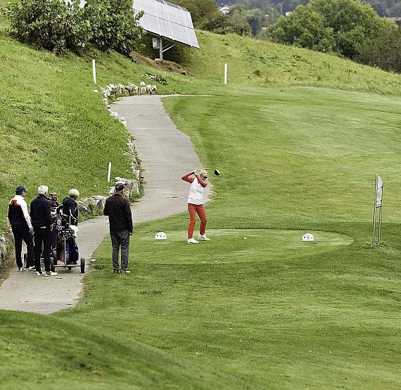 Eine Golferin schlägt auf einem grünen Golfplatz ab. Links stehen Zuschauer, einige mit Golfschlägern, die die Szene beobachten. Im Hintergrund Hügel.
