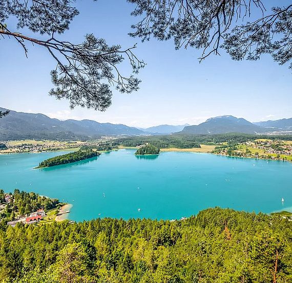 View of a clear blue lake surrounded by trees and mountains under a bright sky, photographed from a high vantage point.