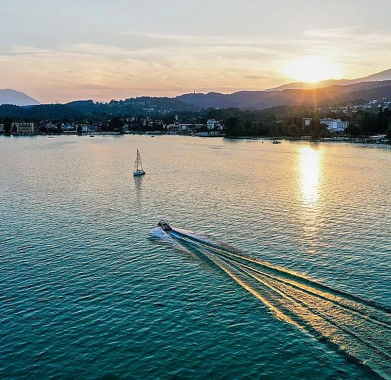 A scenic lake view at sunset with a motorboat leaving a trail, surrounded by distant hills and a glowing sun reflecting on the water's surface.