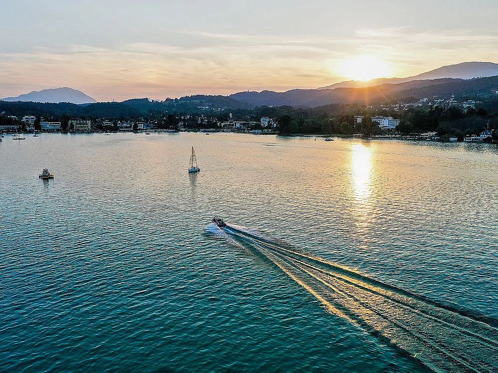 Ein Motorboot zieht Wellen auf einem ruhigen See bei Sonnenuntergang. Im Hintergrund sind Berge und eine Stadt zu sehen, während die Sonne über dem Horizont steht.