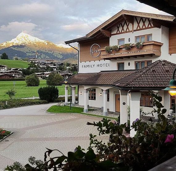 A charming hotel with Alpine architecture sits in a valley with a snow-capped mountain in the distance, surrounded by green fields and a cloudy sky.