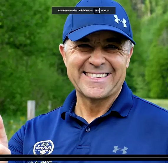 A smiling man in a blue cap and shirt gives a thumbs up in a grassy outdoor setting with trees behind.