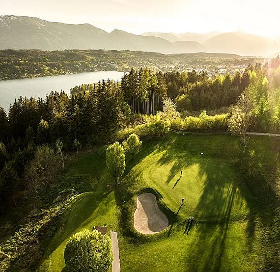 Landschaft mit einem Golfplatz am Waldrand, umgeben von Bäumen und einem See im Hintergrund bei Sonnenuntergang. Die Berge sind in der Ferne sichtbar.