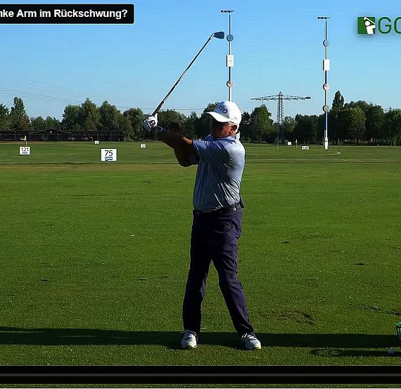 A golfer swings a club on a practice range with a bag beside him and green trees in the background.
