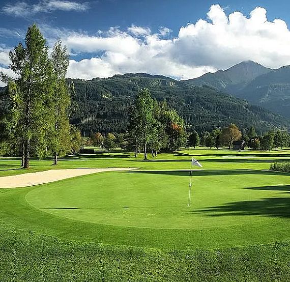 A golf course with lush greens, trees, a sand bunker, and mountains in the background under a blue sky.