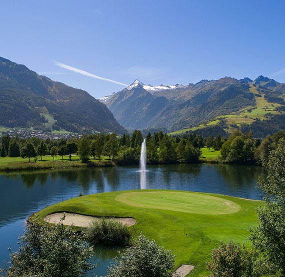 A golf course with a central green, surrounded by a lake with a fountain, set against mountains under a clear blue sky.