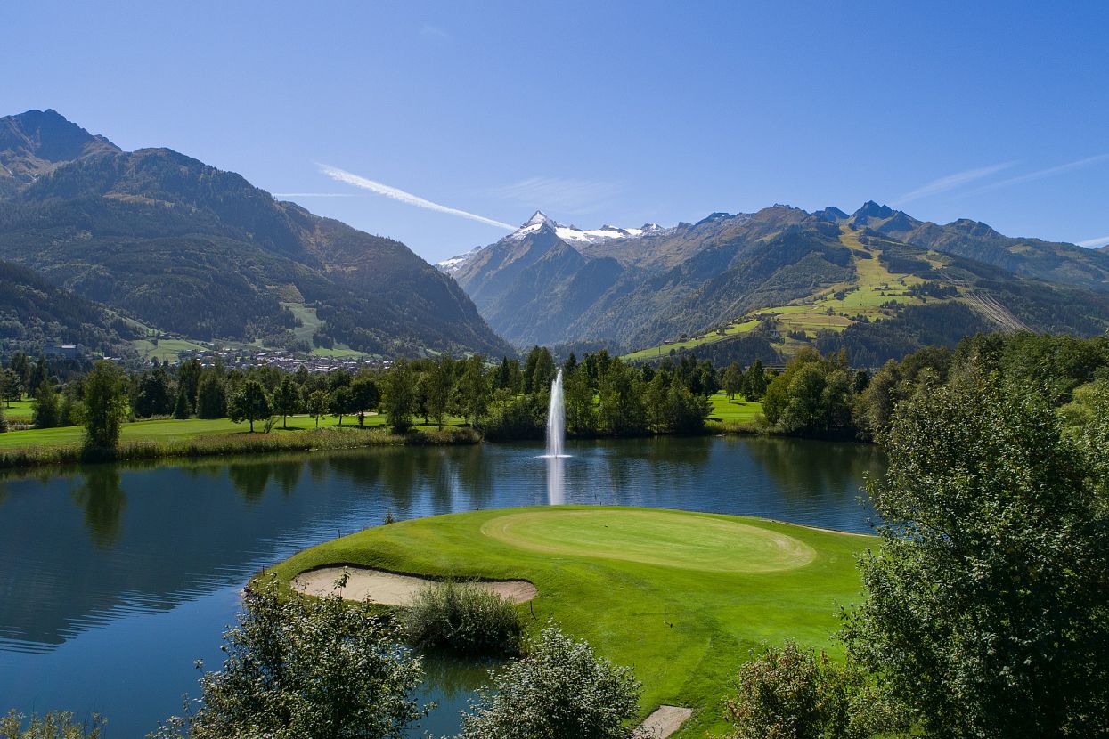 Malersiche Landschaft mit einem Golfplatz im Vordergrund, umgeben von einem See und hohen Bergen im Hintergrund unter klarem blauem Himmel.