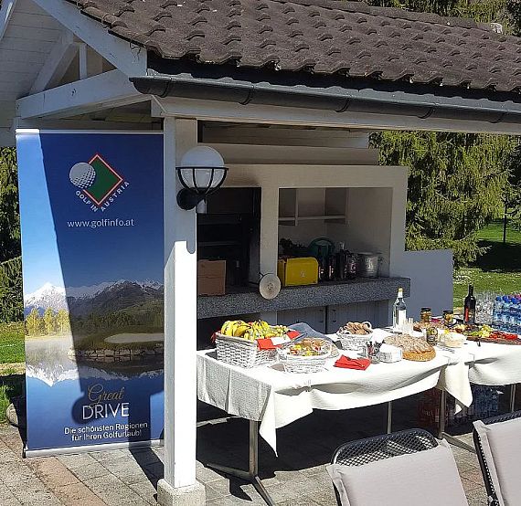 Outdoor pavilion with a buffet table full of food and drinks, surrounded by greenery and a blue sky.