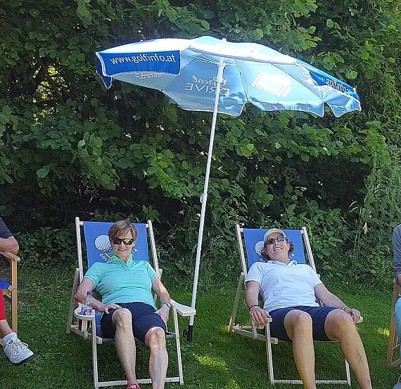 Four people relax on deck chairs in a garden under a blue and white umbrella, surrounded by green foliage, enjoying a sunny day.