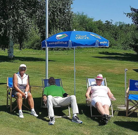 Three adults sit under a blue umbrella on foldable chairs in a sunny, green park. They appear relaxed, surrounded by green grass and trees.