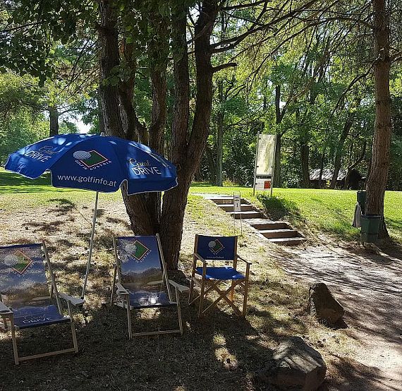 Three blue chairs with umbrellas in a park, surrounded by trees and greenery, with a pathway and stairs nearby, creating a tranquil outdoor setting.