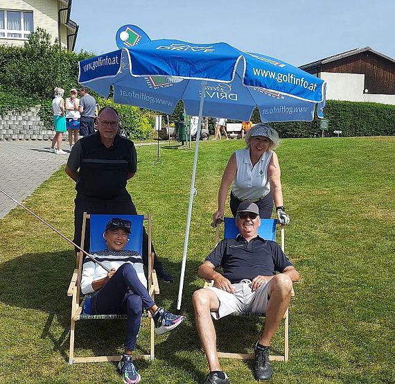Four people at a golf course; two seated in deck chairs under a blue umbrella, two standing behind with golf clubs, smiling amidst a sunny landscape.