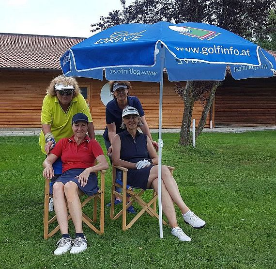Four people at a golf course, two sitting under a blue umbrella and two standing behind them, with a wooden building and trees in the background.