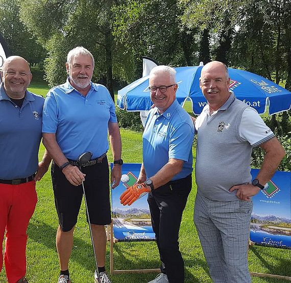 Vier Männer in bunter Golfkleidung stehen lächelnd auf einem grünen Golfplatz. Im Hintergrund sind Sonnenschirme und Bäume zu sehen. Entspannte Atmosphäre.