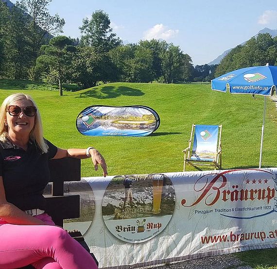 A woman in sunglasses and pink pants sits on a bench at a golf course with green grass, chairs, and banners on a sunny day.