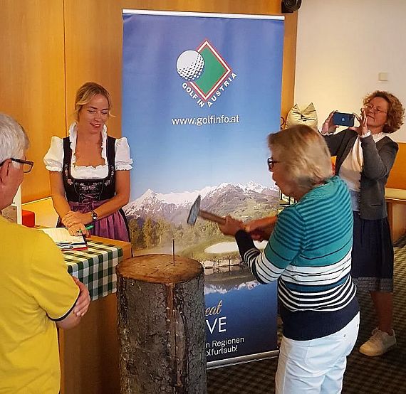 Axe-throwing event indoors with participants and spectators. A woman in a dirndl assists while others watch and photograph. Alpine theme decor.