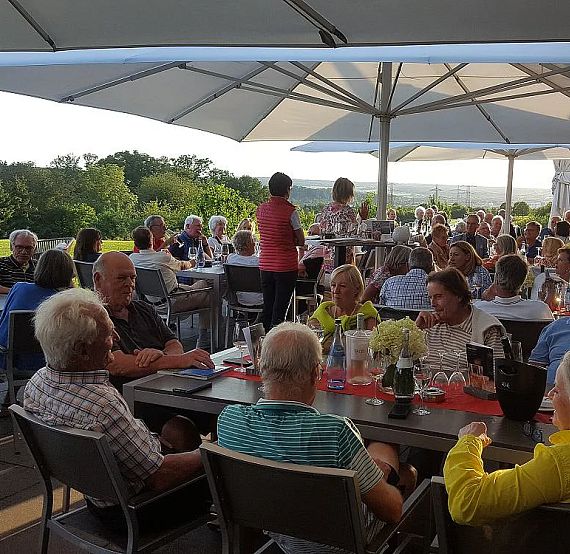 People gather at outdoor tables under umbrellas, enjoying a sunny day and lush greenery. The scene is lively with a relaxed, social atmosphere.