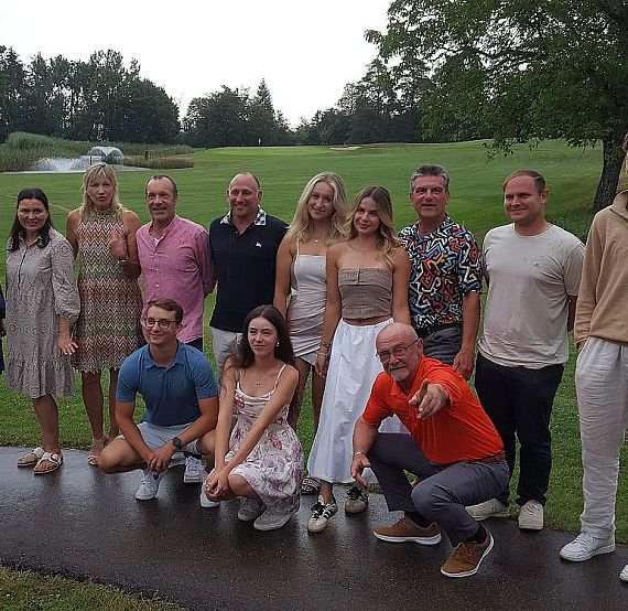 A diverse group of men and women outdoors on a golf course, posing together with trees and green grass in the background, dressed in casual and colorful attire.