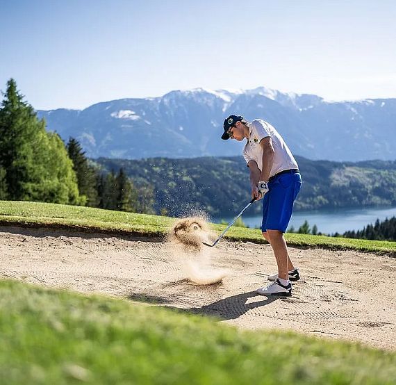 A golfer takes a swing in a sand bunker on a sunny day, surrounded by trees, a lake, and mountains.