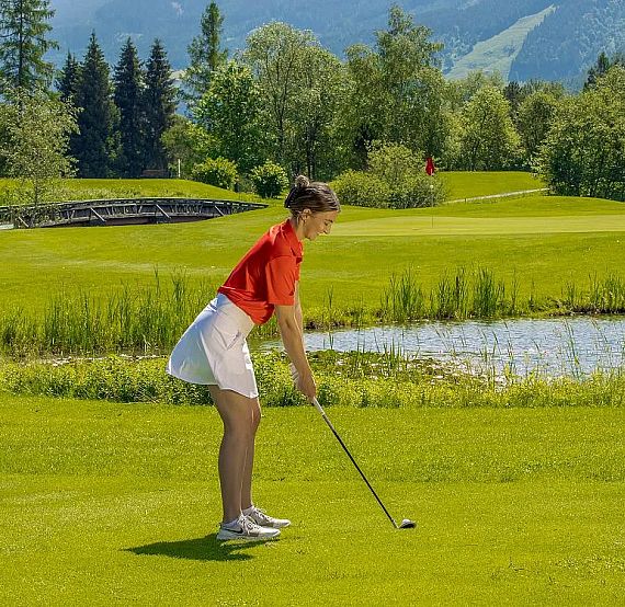 A person in a red shirt and white skirt prepares to hit a golf ball on a lush green course with trees and hills in the background.