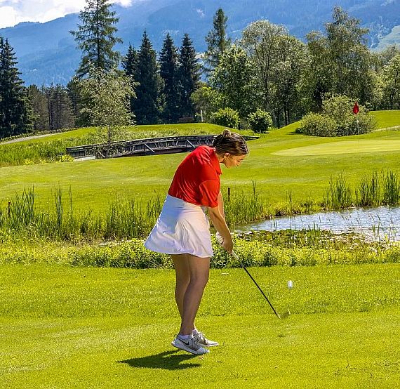 Person golfing on a vibrant green course with mountains, trees, and a pond in the background, under a partly cloudy sky.