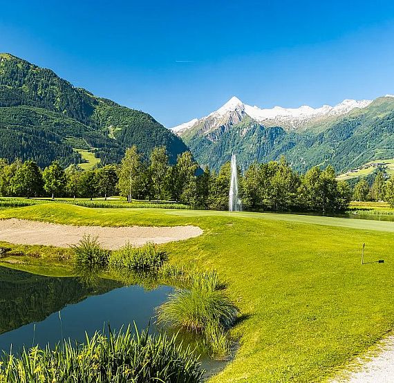 Golf course nestled in lush mountains, featuring a sand trap and pond under a blue sky with a mountain backdrop.