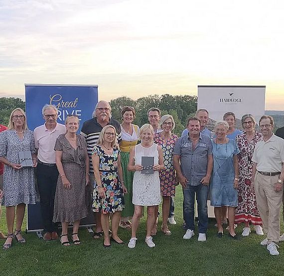 A group of around 20 people smiling and holding trophies on a golf course at sunset, wearing casual clothing with banners in the background.