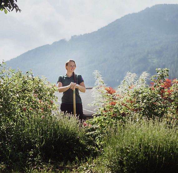 Person holding a shovel in a garden with lush greenery and colorful flowers, set against a mountain backdrop under a cloudy sky.