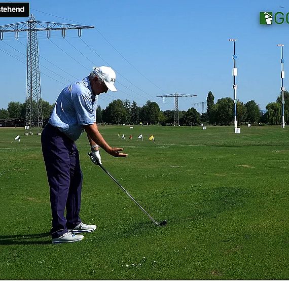 A golfer on a grassy field prepares to swing a club, with utility poles and scattered golf balls in the background under a clear blue sky.