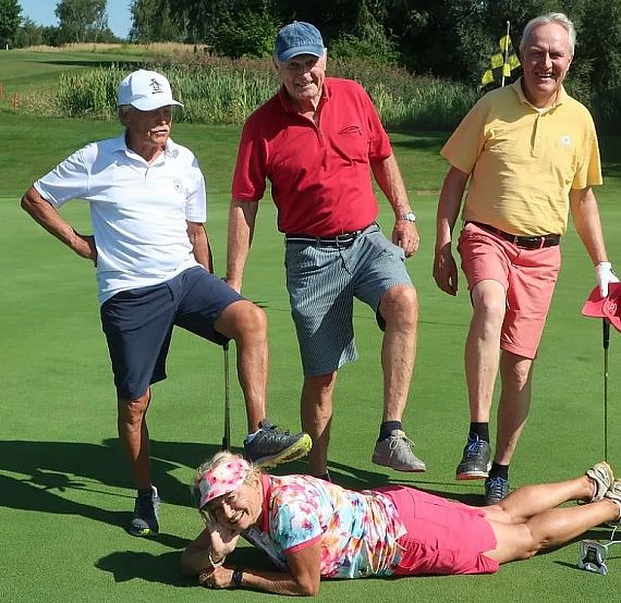 Four people are on a golf course. Three are standing with one foot on a lying person, who is smiling. They appear to be in a playful mood under the sun.