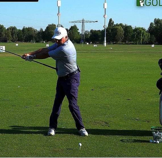 Golfer practicing a swing on a green field, wearing a cap and holding a golf club. A bag and balls are on the grass nearby, with trees in the background.