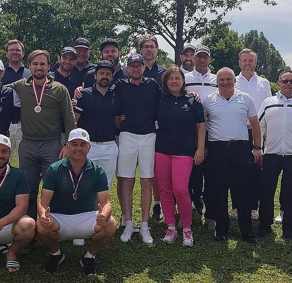 A group of people standing on grass, wearing medals and casual clothing, with trees in the background, possibly at a golf event.