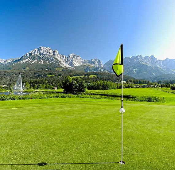 A golf course with a flag on the green, lush greenery, and mountains in the background under a clear sky.