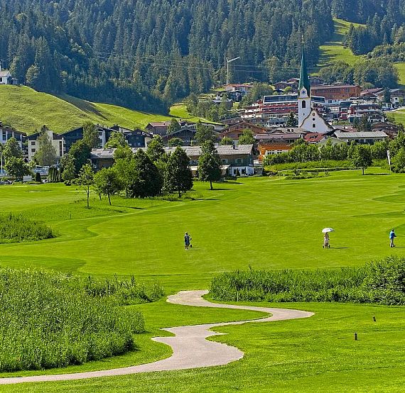 Lush green golf course with players, surrounded by hills and a village with a church steeple, under a blue sky in a mountainous area.