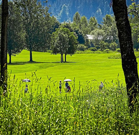 People walking on a golf course surrounded by green grass and trees, with mountains in the background under a clear blue sky.