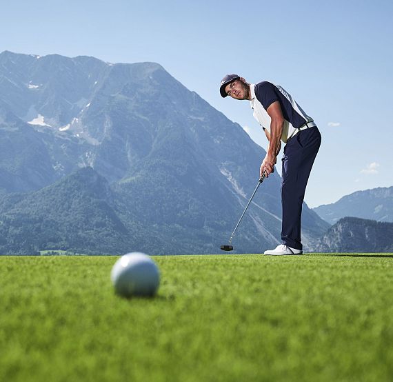 Ein Mann in Sportkleidung steht auf einem Golfplatz mit grüner Wiese und bergiger Landschaft im Hintergrund und bereitet sich darauf vor, einen Golfball zu schlagen.
