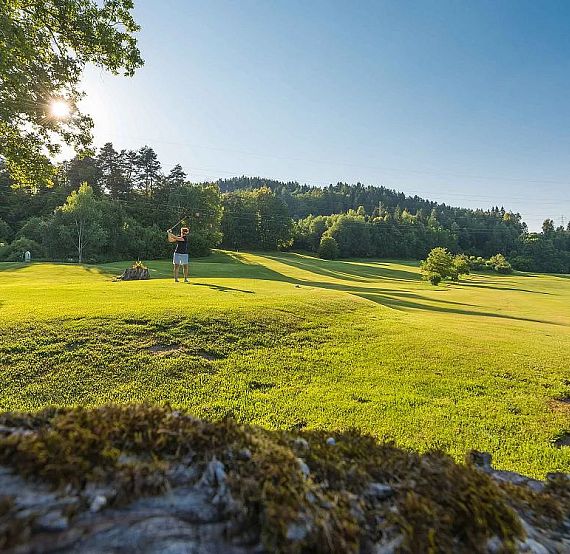 Lush golf course with two distant players under a clear blue sky, surrounded by trees and sunlight casting shadows on the green.