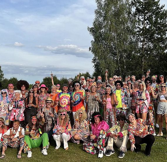 A joyful crowd dressed in colorful 70s attire is gathered outdoors on a grassy field, with trees in the background under a partly cloudy sky.