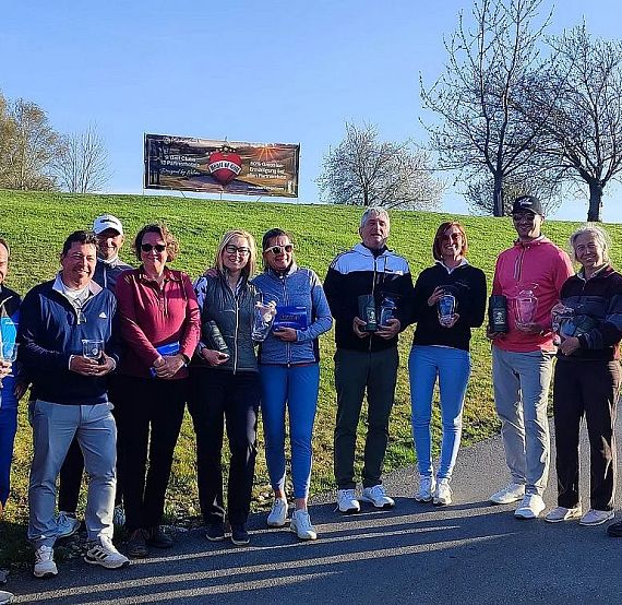 A group of people standing outdoors holding trophies on a sunny day. They are on a pavement with green grass and trees in the background.
